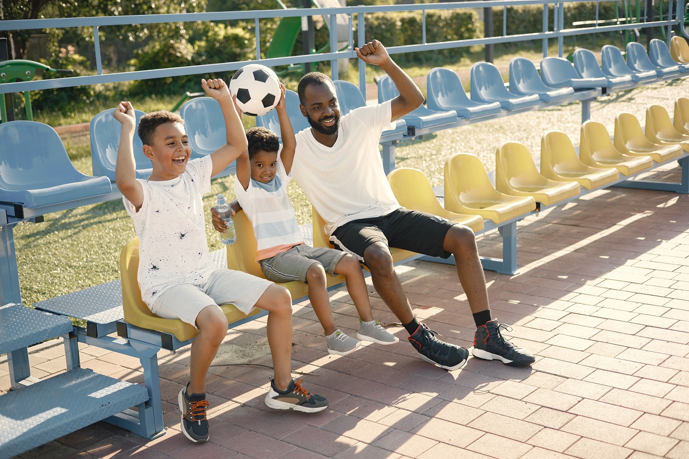 Excited father and two his sons watching football game on stadium and cheering victory. African man and two his multiracional sons wearing white t-shirts. Boy holding a ball.